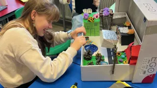 Zwei Mädchen im Klassenzimmer, eine benutzt ein Tablet und die andere dekoriert ein kleines Hausmodell in einem Karton.