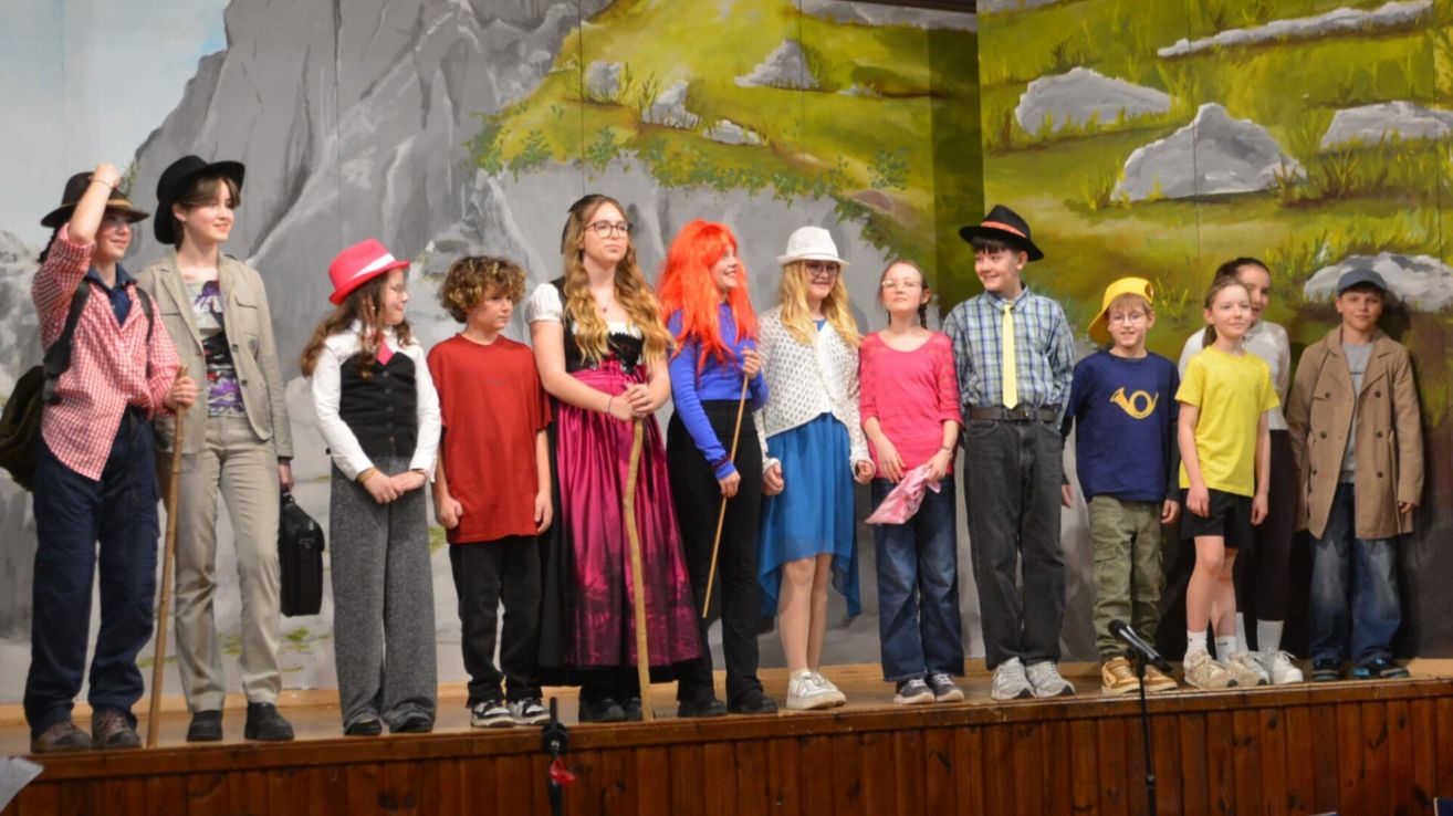 A group of children dressed in various costumes stand on a stage with a painted backdrop behind them. Some hold props.