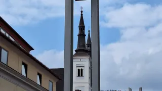 Ein hoher Kirchturm steht gegen einen bewölkten Himmel, umgeben von Gebäuden mit roten Dächern, aus einer niedrigen Perspektive sichtbar.
