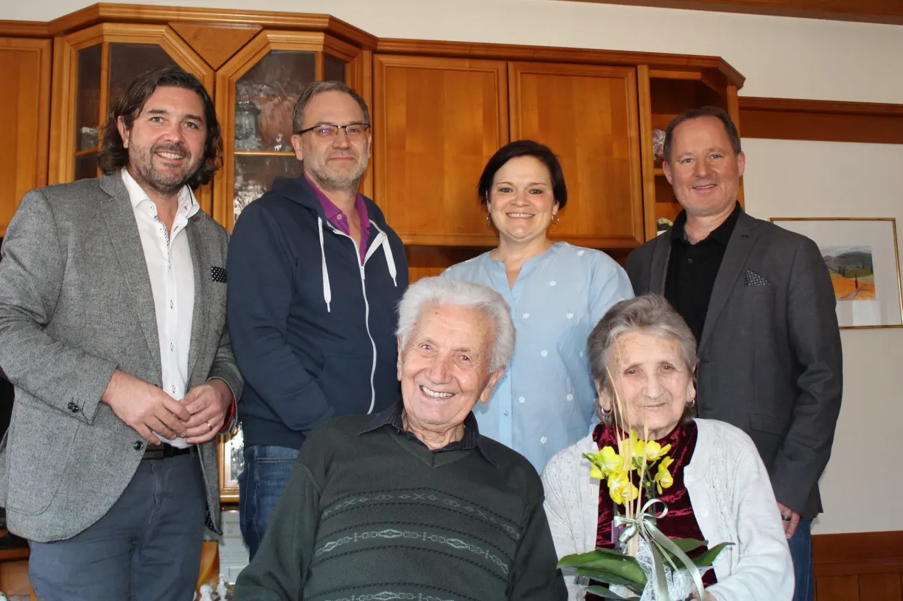 Eine Gruppe von Menschen, darunter ein älteres Paar, steht in einem Raum mit hölzernen Schränken im Hintergrund. Das ältere Paar lächelt und hält einen Strauß Blumen.