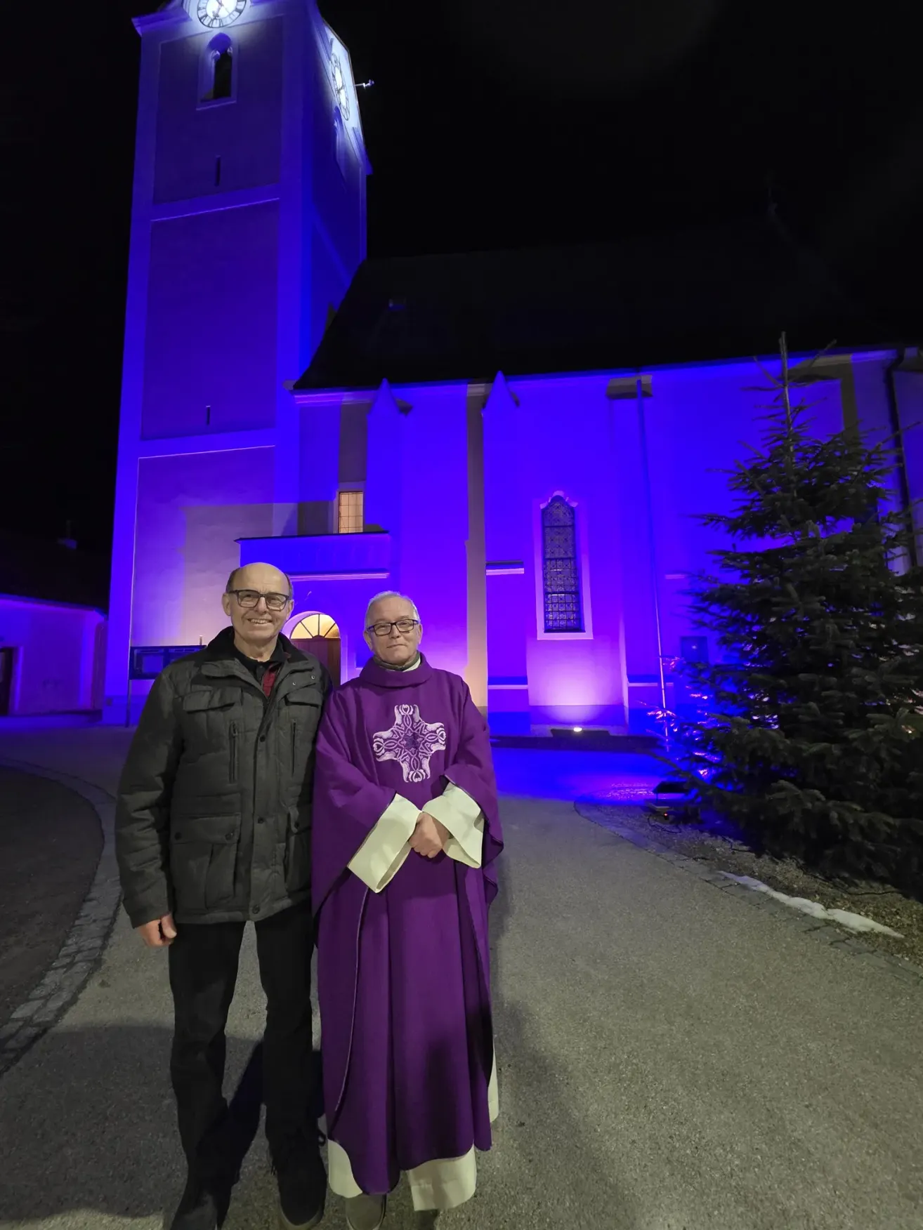 Zwei Männer stehen vor einer Kirche mit blauer Beleuchtung, einer mit Brille und einem schwarzen Mantel, der andere in einem violetten Gewand.