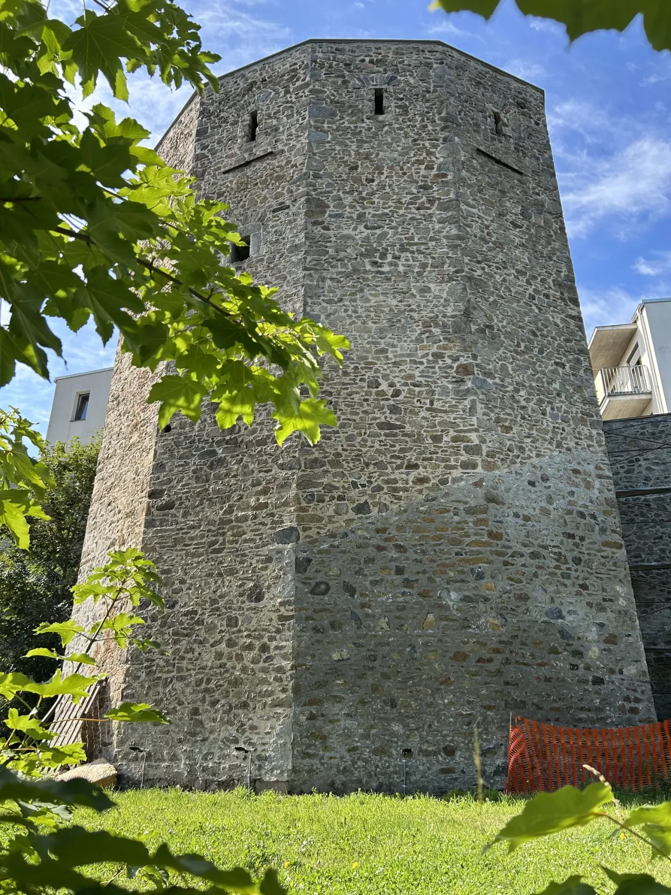 A tall stone tower stands prominently against a blue sky, surrounded by lush greenery. A modern building with a balcony is visible to the right.