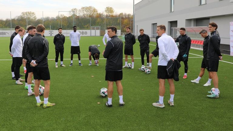 A group of soccer players and coaches on a field, one player bending down to pick up a ball. In the background, a building with a sponsor logo.