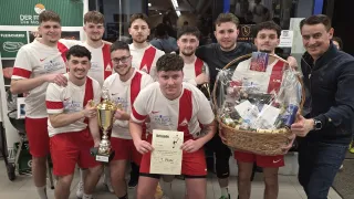 Eine Gruppe von Männern in passenden Uniformen posiert für ein Foto mit einem Pokal und einem Korb voller Geschenke. Sie scheinen einen Sieg zu feiern.