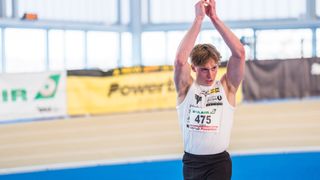 Ein junger männlicher Athlet mit der Nummer 475 auf seinem Shirt streckt seine Arme über dem Kopf. Er trägt ein weißes Tanktop und schwarze Shorts. Hinter ihm ist ein gelber Banner mit dem Wort 'Power'.