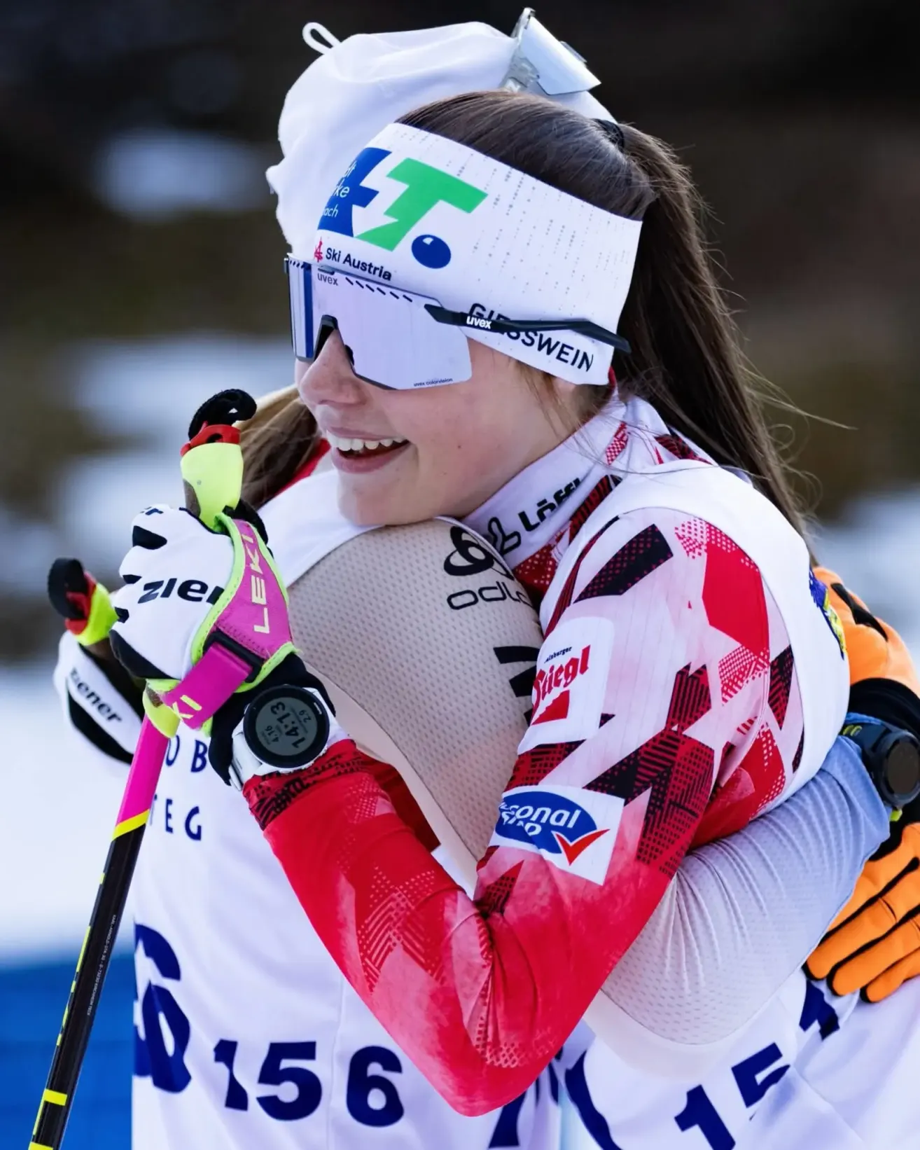 Eine lächelnde Skifahrerin umarmt eine andere Skifahrerin in einem verschneiten Außenbereich, beide tragen Schutzbrillen, Handschuhe und Uhren. Die Skifahrerin im Vordergrund trägt ein weißes und rotes Outfit mit Logos.