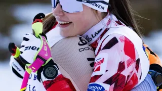 Eine lächelnde Skifahrerin umarmt eine andere Skifahrerin in einem verschneiten Außenbereich, beide tragen Schutzbrillen, Handschuhe und Uhren. Die Skifahrerin im Vordergrund trägt ein weißes und rotes Outfit mit Logos.