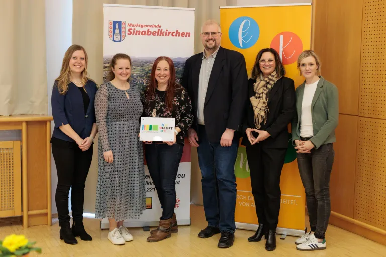 Eine Gruppe von Menschen steht lächelnd vor einem Banner. Eine Frau hält ein Zertifikat. Das Banner trägt den Schriftzug 'Marktgemeinde Sinabelkirchen'. Dahinter befindet sich ein weiteres Banner mit 'E' und 'K'. Die Gruppe besteht aus vier Frauen und einem Mann.