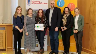 Eine Gruppe von Menschen steht lächelnd vor einem Banner. Eine Frau hält ein Zertifikat. Das Banner trägt den Schriftzug 'Marktgemeinde Sinabelkirchen'. Dahinter befindet sich ein weiteres Banner mit 'E' und 'K'. Die Gruppe besteht aus vier Frauen und einem Mann.