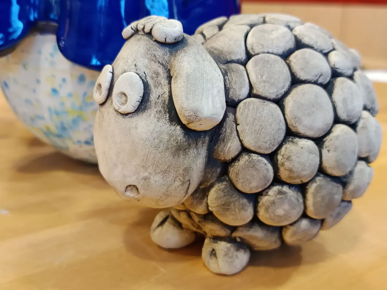 Eine Nahaufnahme einer Keramik-Schafsfigur. Das Schaf hat einen felsartigen Körper, einen weißen Kopf und weiße Augen. Es steht auf einer Holzoberfläche mit einem blauen Gefäß dahinter.