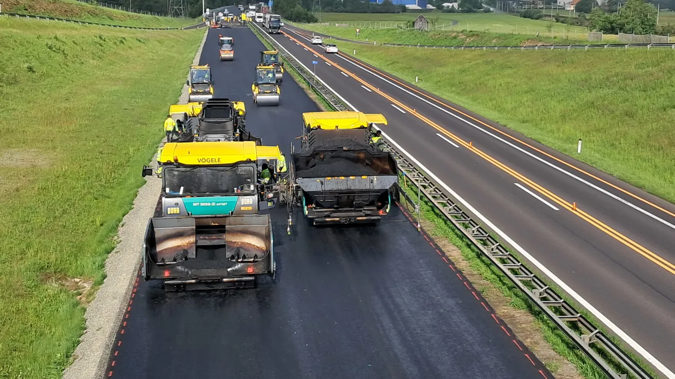 Eine Straßenbaubrigade nutzt schwere Maschinen, um neuen Asphalt auf einer Autobahn zu verlegen. Mehrere gelbe und schwarze Fahrzeuge sind bei der Arbeit auf der Straße zu sehen.
