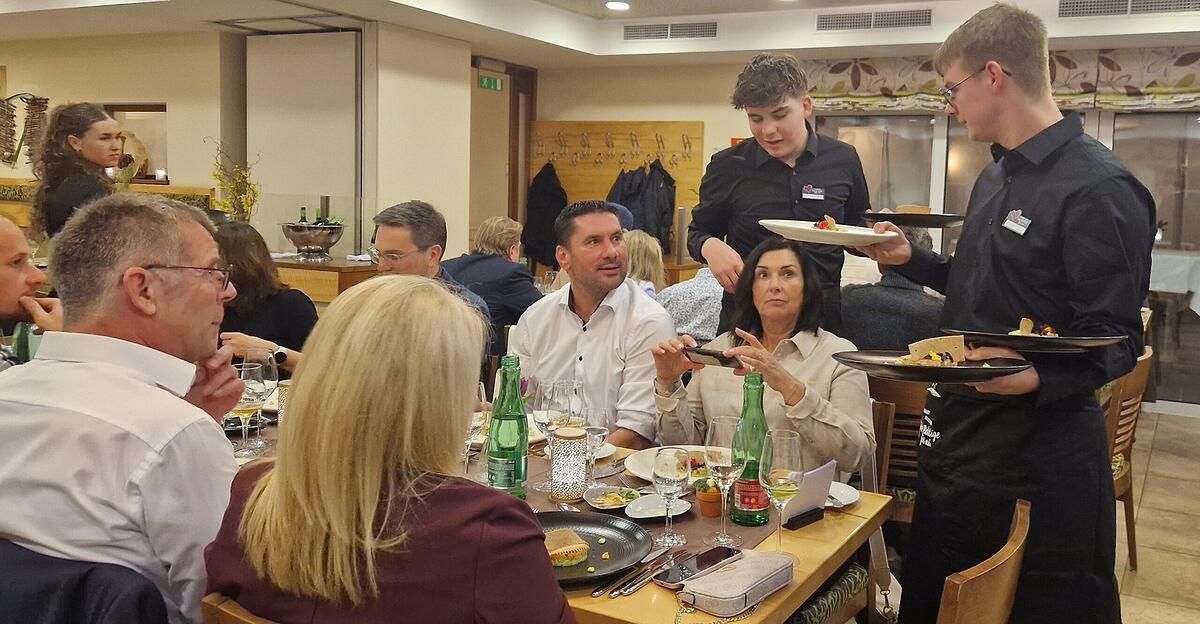 A group of people sit at a table in a restaurant. Two waiters serve them. One woman is taking a picture with her phone.