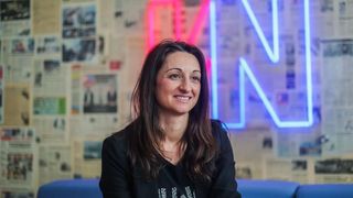 Eine lächelnde Frau mit langen braunen Haaren sitzt auf einem blauen Sofa in einem Büro. Hinter ihr ist eine Wand mit Zeitungen bedeckt und ein rosa und blaues Neonschild mit dem Buchstaben N.