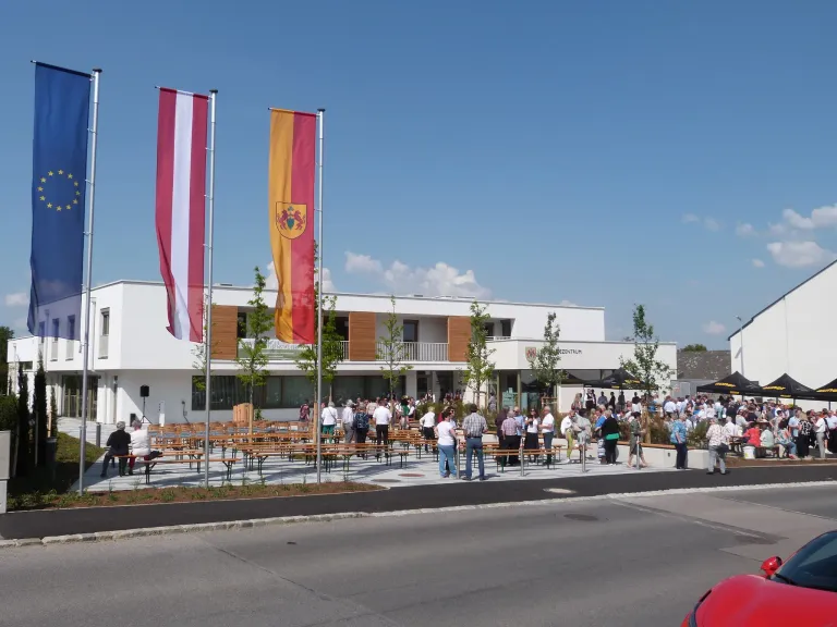 A crowd of people outside a building with flags. Some are sitting on benches while others are walking on the sidewalk. Trees and plants are in front of the building.