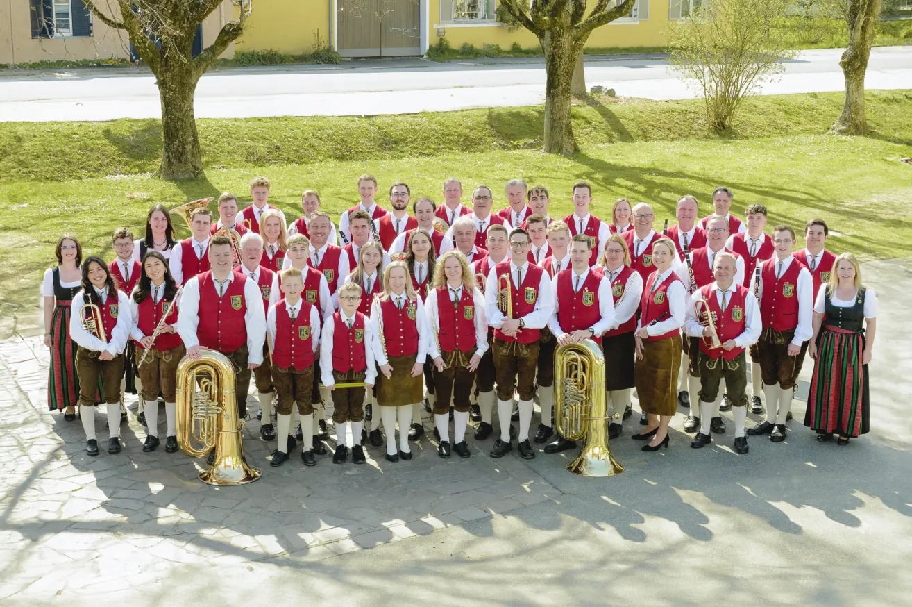 A large group of musicians, both men and women, dressed in traditional Bavarian attire, pose for a photo in a grassy area with trees and a building in the background.