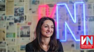 Eine lächelnde Frau mit langen Haaren steht vor einem Hintergrund aus Zeitungsausschnitten und einem Neonschild mit den Buchstaben 'MN'.