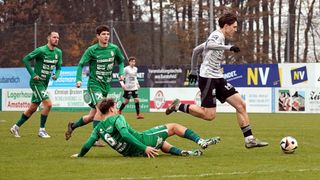 Ein Fußballspiel mit Spielern in grünen und weißen Trikots, ein Spieler rutscht auf dem Rasen, während ein anderer vorbeiläuft. Im Hintergrund befindet sich ein Zaun mit Bannern und Bäumen.