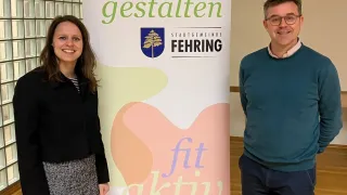 Zwei Personen stehen neben einem Banner mit einer motivierenden Gesundheitsbotschaft, 'fit aktiv gesund,' und dem Logo der Stadtgemeinde Fehring. Die Frau trägt einen schwarzen Mantel und der Mann ein türkisfarbenes Sweatshirt.