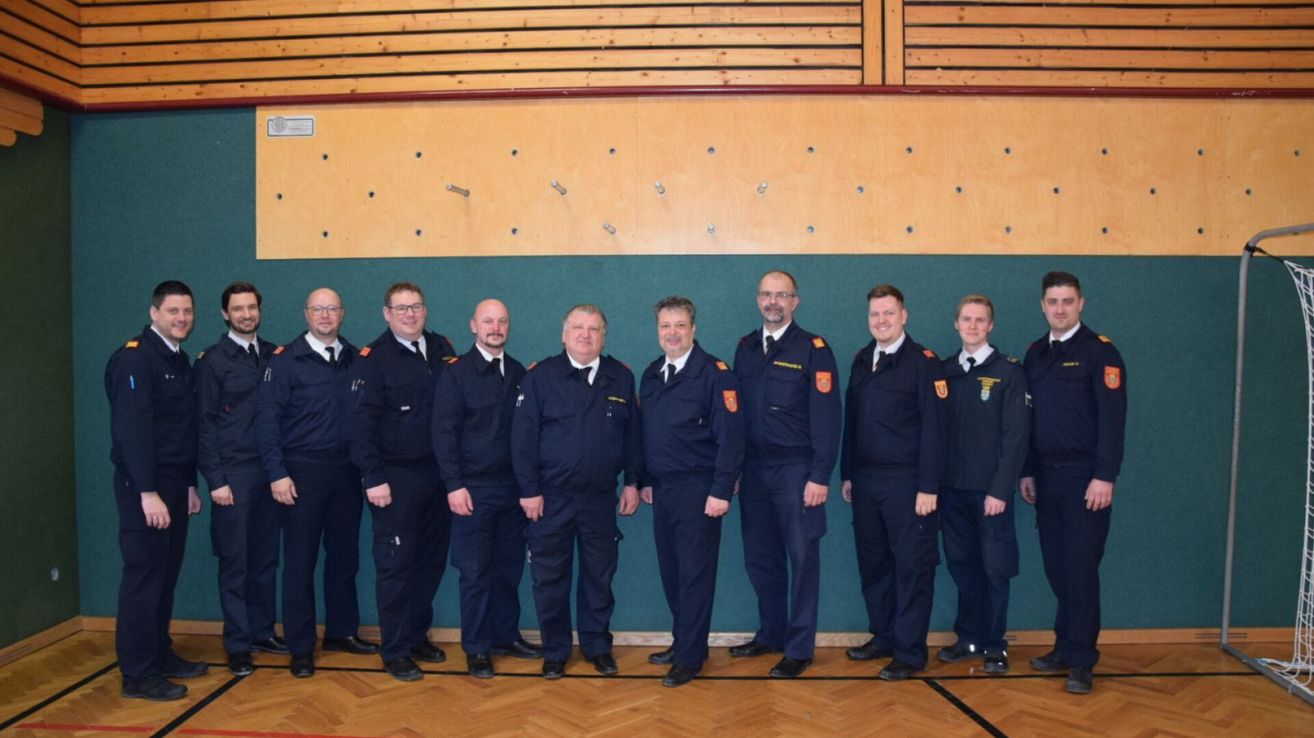 A group of uniformed men stand in a row in front of a green wall with wooden panels above and below, possibly a sports hall. They wear badges on their uniforms.