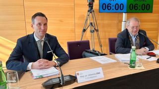 Zwei Männer in Anzügen sitzen in einem Konferenzraum an einem Tisch mit einem digitalen Timer und einem Mikrofon. Papiere und ein Stift liegen auf dem Tisch.