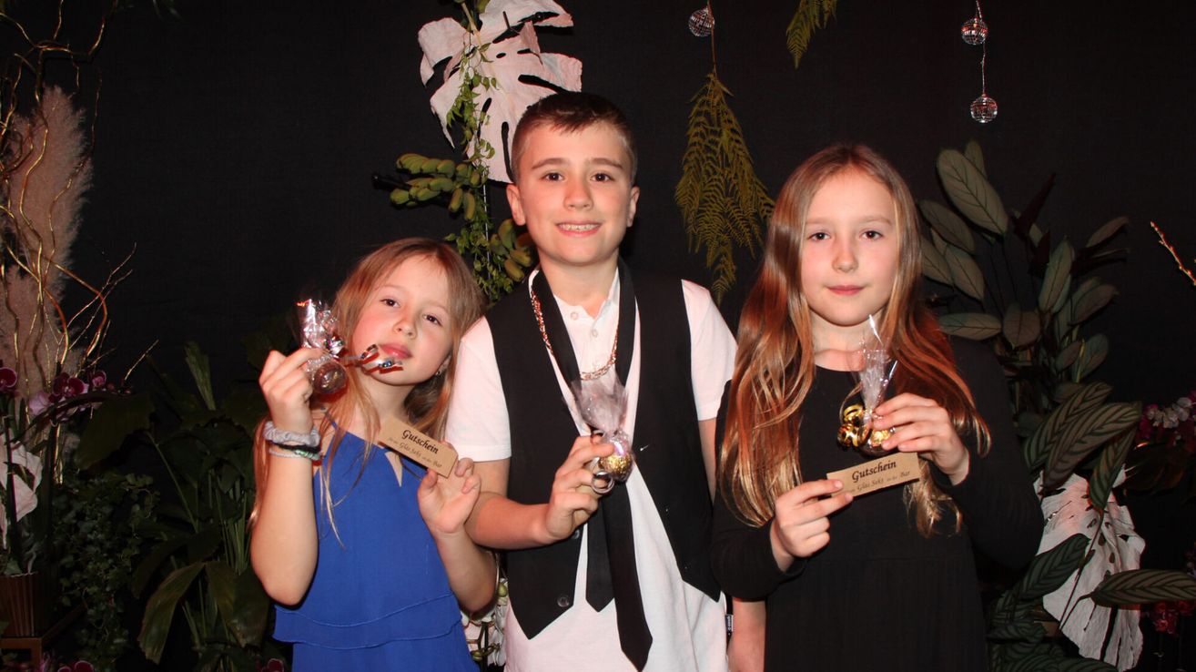 Drei Kinder posieren mit verpackten Geschenken; ein Junge in einem schwarzen Weste und Krawatte, ein Mädchen in Blau und ein weiteres in Schwarz. Sie stehen vor einem Hintergrund mit grünen Blättern.