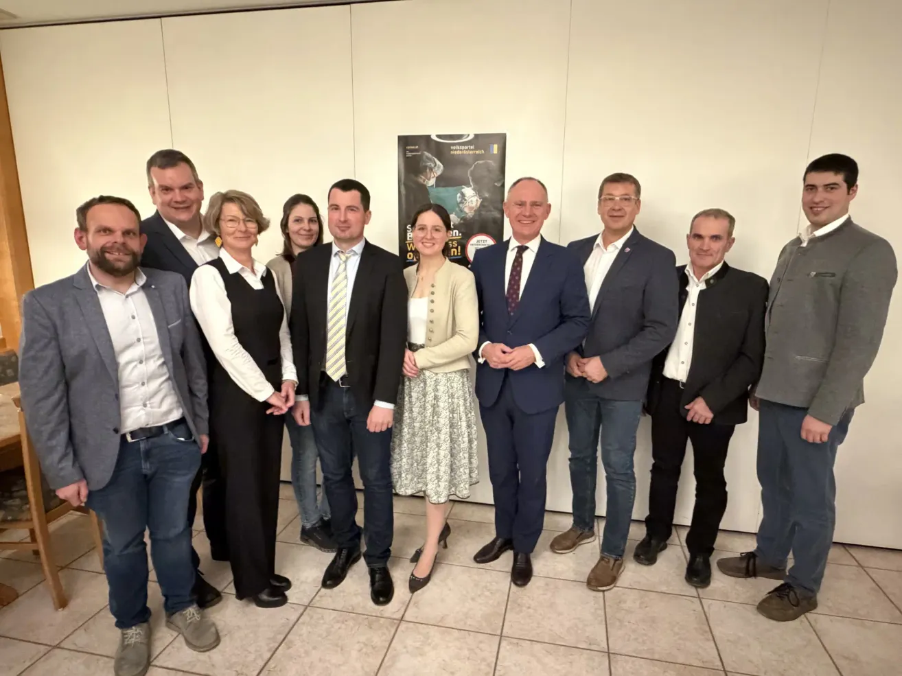 Eine Gruppe von Menschen in formeller Kleidung posiert für ein Foto in einem Raum, mit einem Plakat im Hintergrund.