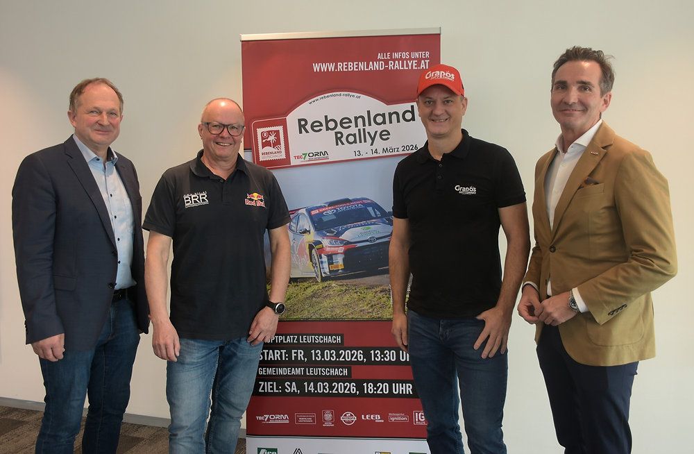 Vier Männer stehen neben einem Plakat für eine Rallye, in Freizeitkleidung. Das Plakat zeigt ein Rallye-Auto und den Namen der Veranstaltung 'Rebenland Rallye' mit den Daten.