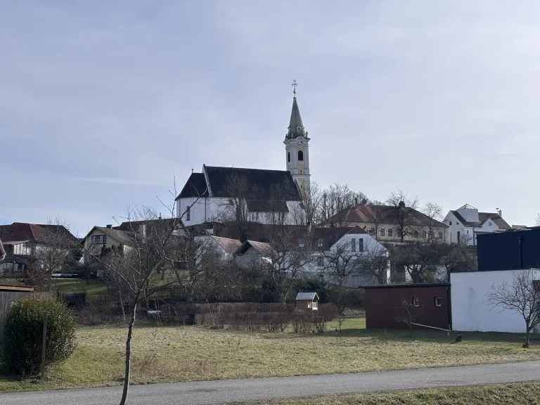 A village with a large church in the center. The church has a steeple and a clock tower. There are houses and buildings around the church. The sky is cloudy.
