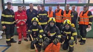 Eine Gruppe Feuerwehrleute in Uniformen und Helmen, einige mit Werkzeugen, posiert für ein Foto in einem Gebäude. Sie stehen vor einem Glasfenster mit Reflexionen.
