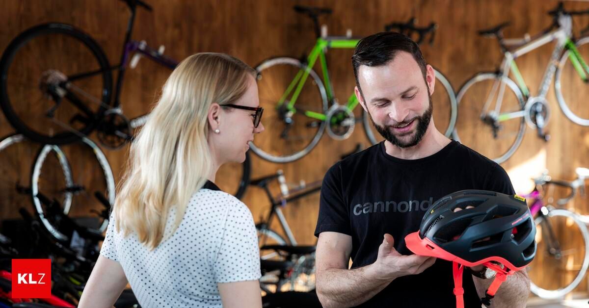 Ein Mann in einem schwarzen Cannondale-Shirt hält einen Helm, während eine Frau mit Brille zusieht. Sie stehen vor einer Wand mit mehreren aufgehängten Fahrrädern.