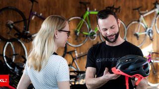 Ein Mann in einem schwarzen Cannondale-Shirt hält einen Helm, während eine Frau mit Brille zusieht. Sie stehen vor einer Wand mit mehreren aufgehängten Fahrrädern.