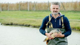 Ein Mann in einem blauen Pullover und grüner Regenkleidung hält einen großen Fisch am See. Er lächelt, steht am Wasser mit einem Grasfeld im Hintergrund.