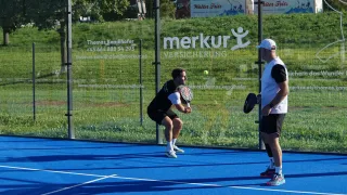 Zwei Männer spielen Tennis auf einem blauen Platz. Einer bereitet sich darauf vor, einen Ball zu schlagen, während der andere zuschaut. Ein Glaszaun mit einem grünen Feld und Bäumen dahinter ist sichtbar.