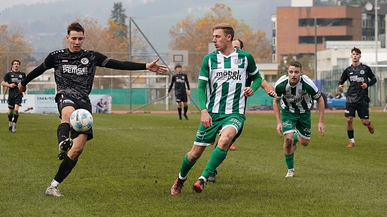 Ein Fußballspieler in einer grünen Uniform läuft mit einem Ball auf das Tor zu, während zwei Spieler in schwarzen Trikots ihm folgen. Das Feld ist von einem Zaun mit Bäumen und Gebäuden umgeben.