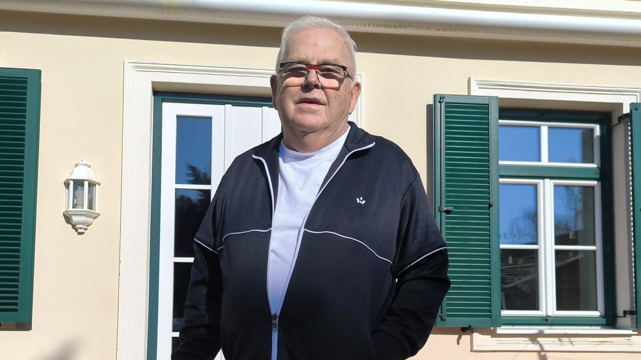 Ein älterer Mann mit Brille und schwarzer Jacke steht vor einem beigen Haus mit grünen Fensterläden und einer weißen Tür.