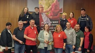 Eine Gruppe von Menschen steht vor einem Banner, das 'Freien-Service' mit einer Kontaktnummer bewirbt. Eine Frau hält ein Werkzeug. Dahinter zeigt ein Banner einen Mann, der einer älteren Person hilft.