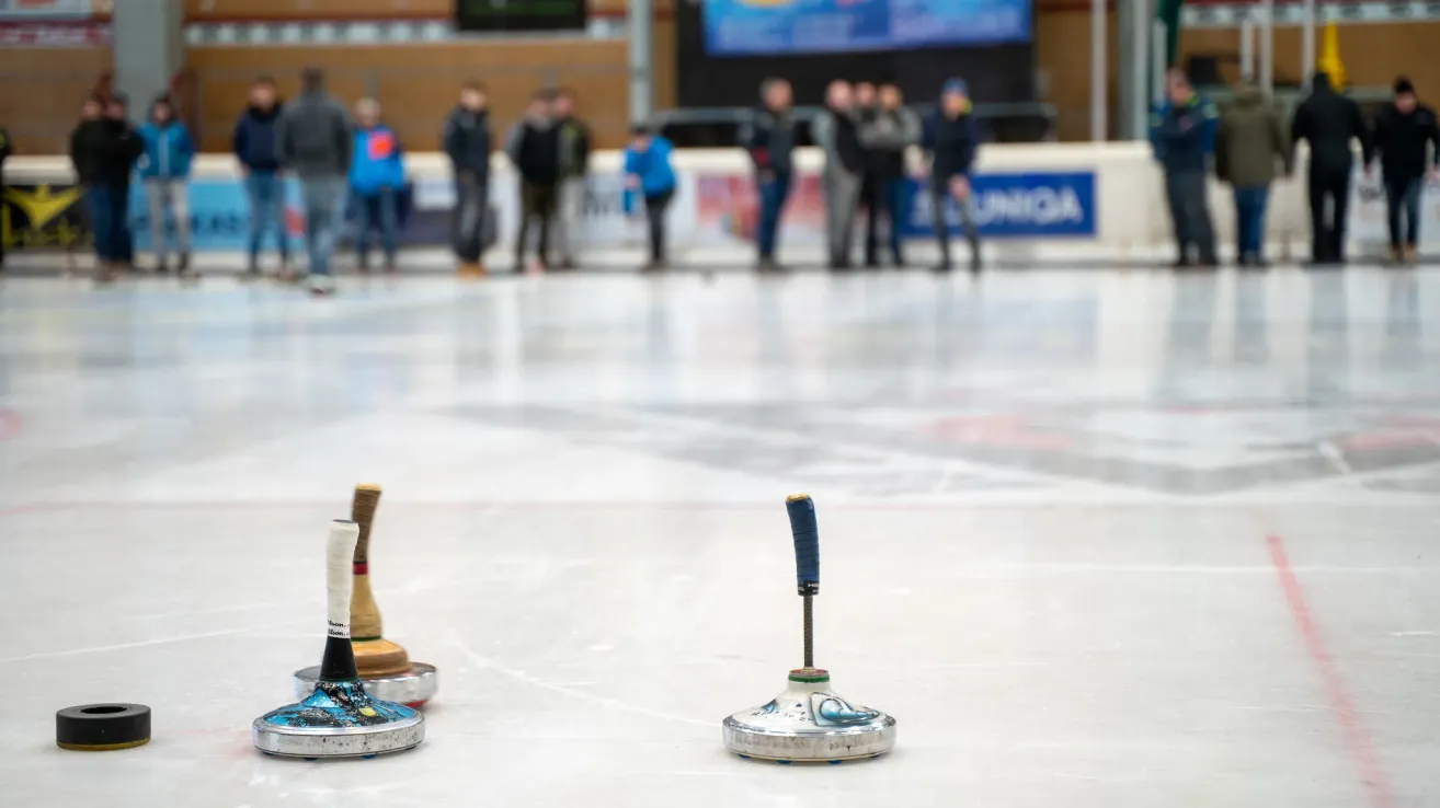 Eine Eisbahn mit Menschen, die herumstehen, zwei Curlern im Vordergrund und einem Schild mit der Aufschrift 'Uniqa'.