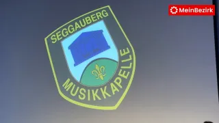 Ein Logo mit einem Schild, das ein Gebäude auf einem Hügel zeigt, umgeben von einem grünen Feld und einem Fleur-de-Lis-Symbol. Die Worte 'SEGGAUBERG MUSIKAPELL' sind in Gelb auf grünem Hintergrund geschrieben.