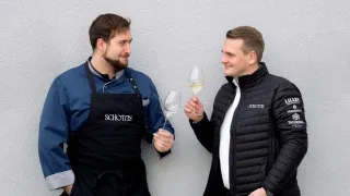 Zwei Männer stehen nebeneinander, beide lächelnd, und halten Weingläser in den Händen. Der Mann auf der linken Seite trägt ein schwarzes Schürzen mit dem Wort Schottis darauf. Der Mann auf der rechten Seite trägt einen schwarzen Mantel.
