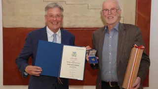 Zwei Männer lächeln, einer hält eine blaue Mappe und der andere eine Medaille. Sie stehen vor einer Wand.