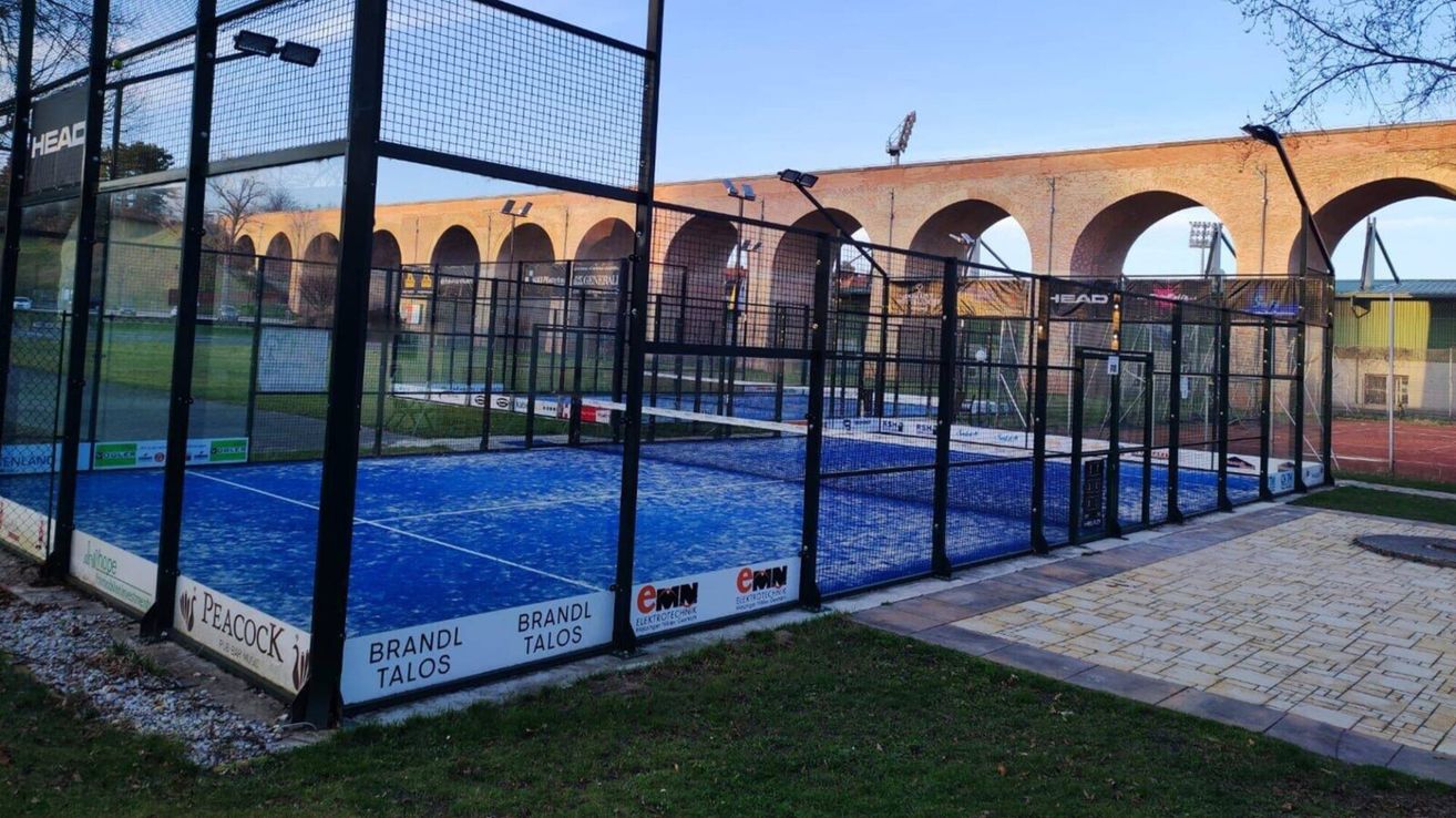 Ein Außentennisplatz mit blauem Boden und schwarzer Umzäunung. Der Zaun hat weißen Text mit 'BRANDL TALOES' und ein Logo mit 'eMMN'. Dahinter befindet sich eine Brücke mit Bögen.