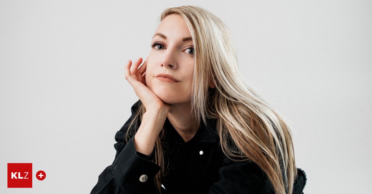 Eine Frau mit blonden Haaren schaut nach oben und stützt ihr Kinn auf ihre Hand, sie trägt eine schwarze Jacke.