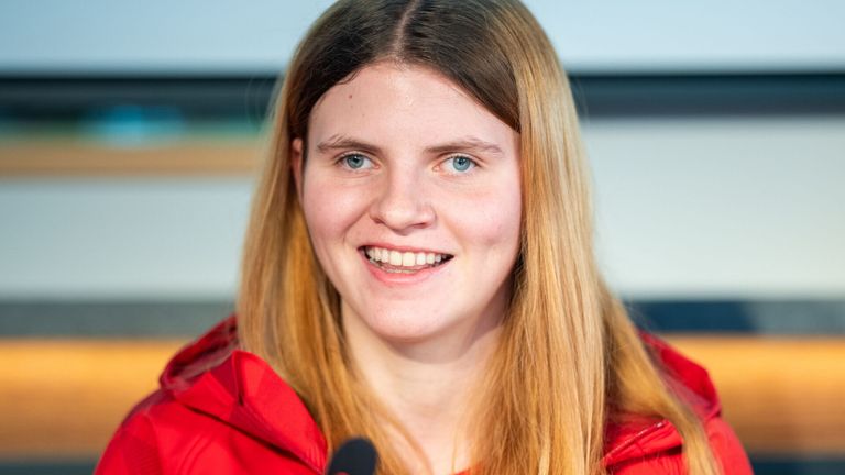 A woman with long blonde hair, wearing a red jacket, smiles for a photo, standing in front of a microphone.
