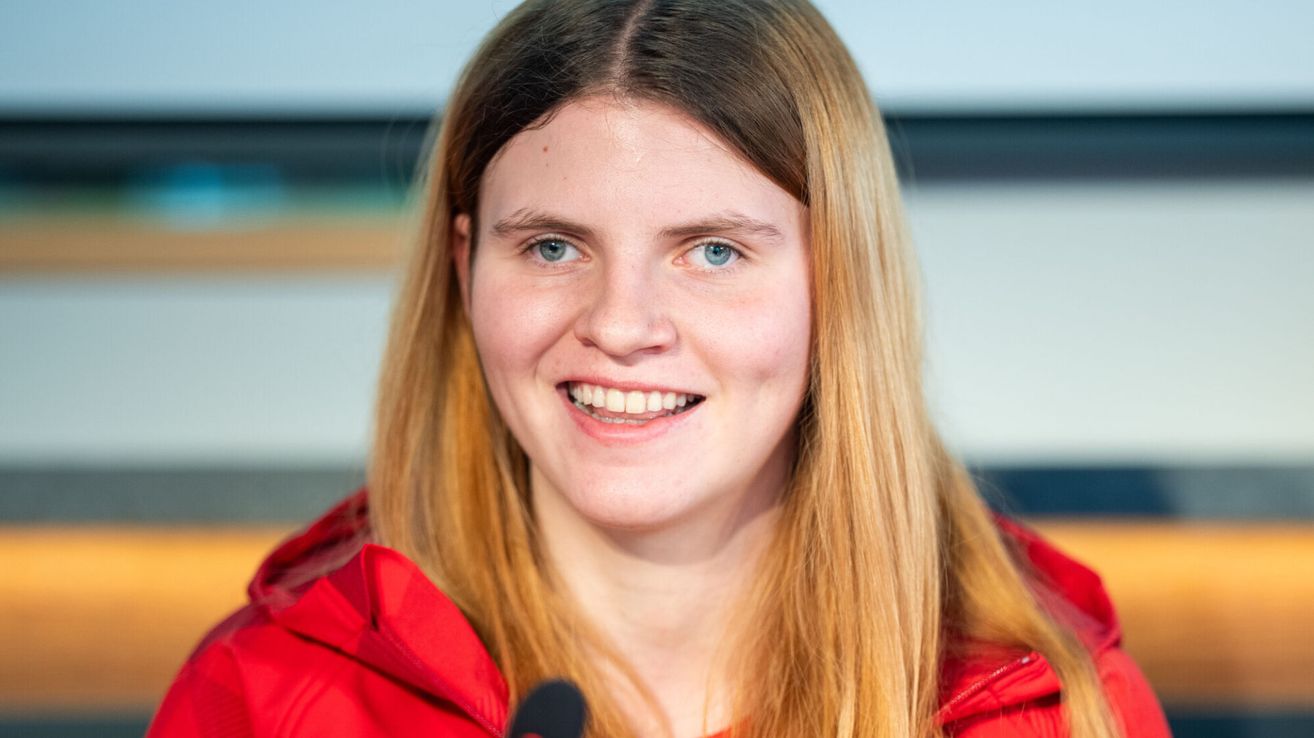 A woman with long blonde hair, wearing a red jacket, smiles for a photo, standing in front of a microphone.