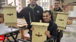 Drei Männer stehen in einer Werkstatt, lächeln und halten Holzkisten. Zwei der Kisten haben Fledermausdesigns darauf. Der Mann in der Mitte ist im Rollstuhl. Hinter ihnen ist ein Feuerlöscher an der Wand montiert.