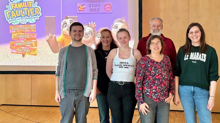 Five individuals stand before a screen showing cartoon characters. A man in gray wears a cardigan, a woman in black has a necklace, and a woman in white wears a tank top. An older man and woman are also present.