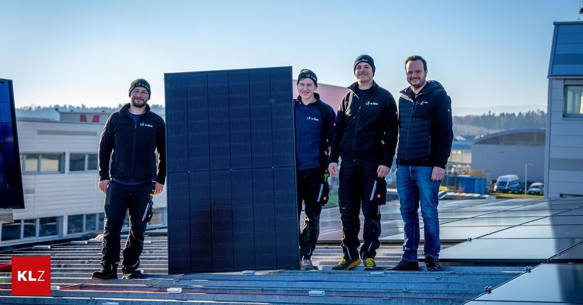 Four men pose with a large solar panel on a rooftop. They are dressed warmly in winter clothing. The solar panel is large and rectangular.