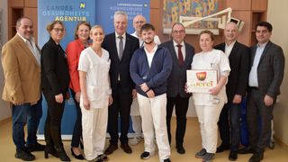 Eine Gruppe von medizinischem Personal und Beamten steht zusammen vor einem Banner. Eine Frau hält eine Box mit der Aufschrift 'merci'.