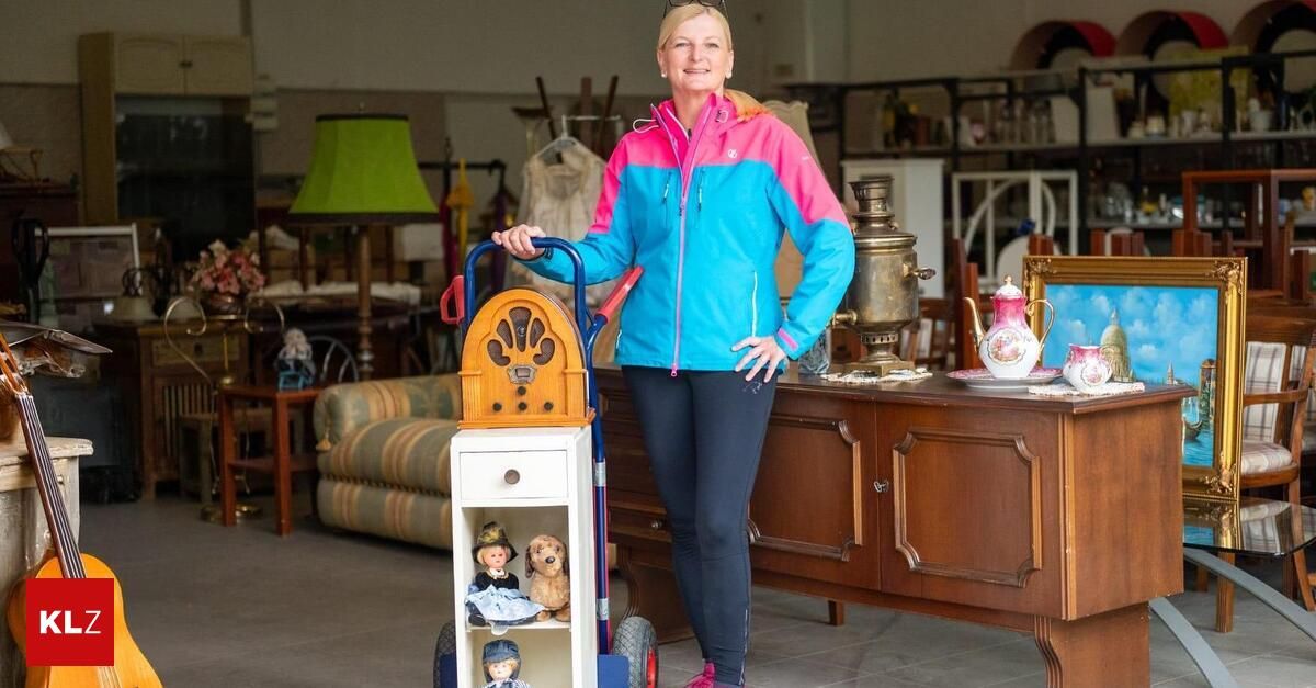 Eine Frau steht in einem Raum mit verschiedenen Möbeln und Gegenständen und hält einen Wagen mit Spielzeug. Sie trägt eine pinke und blaue Jacke und schaut nach vorne.