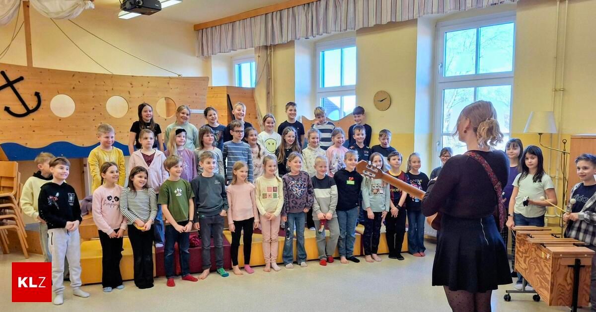 Eine Gruppe von Kindern steht in einem Klassenzimmer, während eine Frau vor ihnen eine Gitarre spielt.